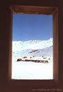 An image of a wooden window frame through which an iron bridge and snow-covered mountains can be seen, in the vicinity of Baños Morales and Baños de la Colina.